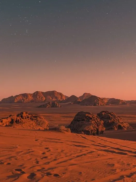 A serene desert landscape at twilight, featuring reddish-orange sand dunes in the foreground, rugged rock formations, and…