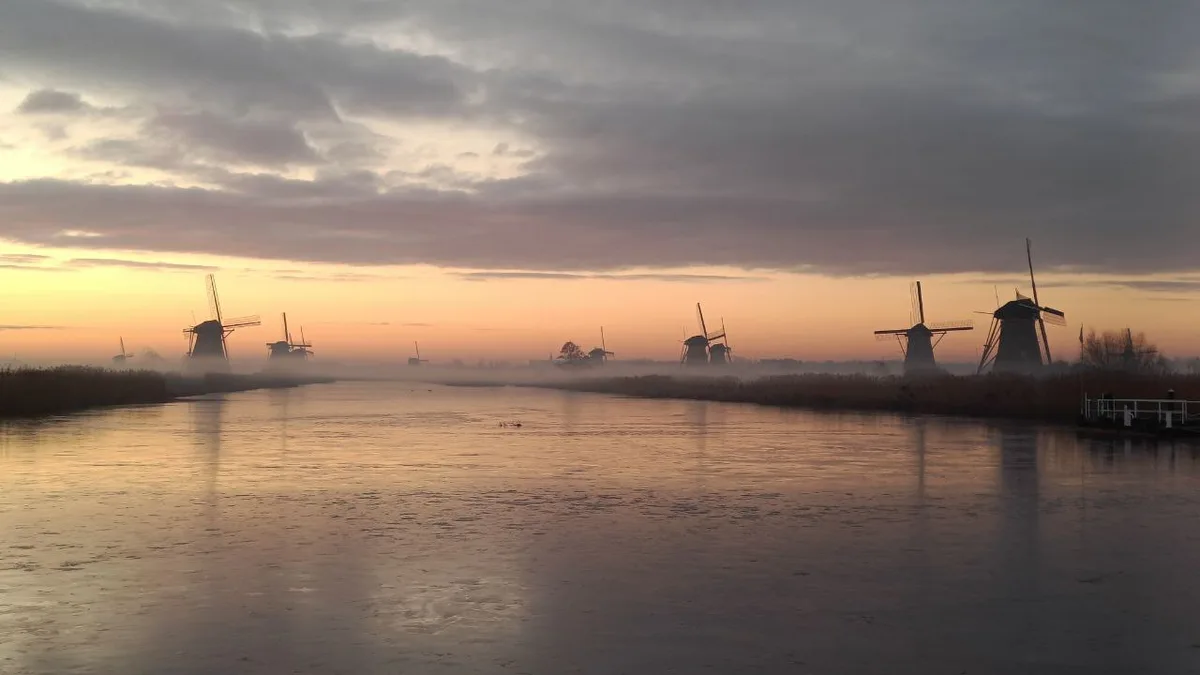 A serene landscape of Dutch windmills silhouetted against a misty, colorful dawn sky over a calm, partially frozen canal…
