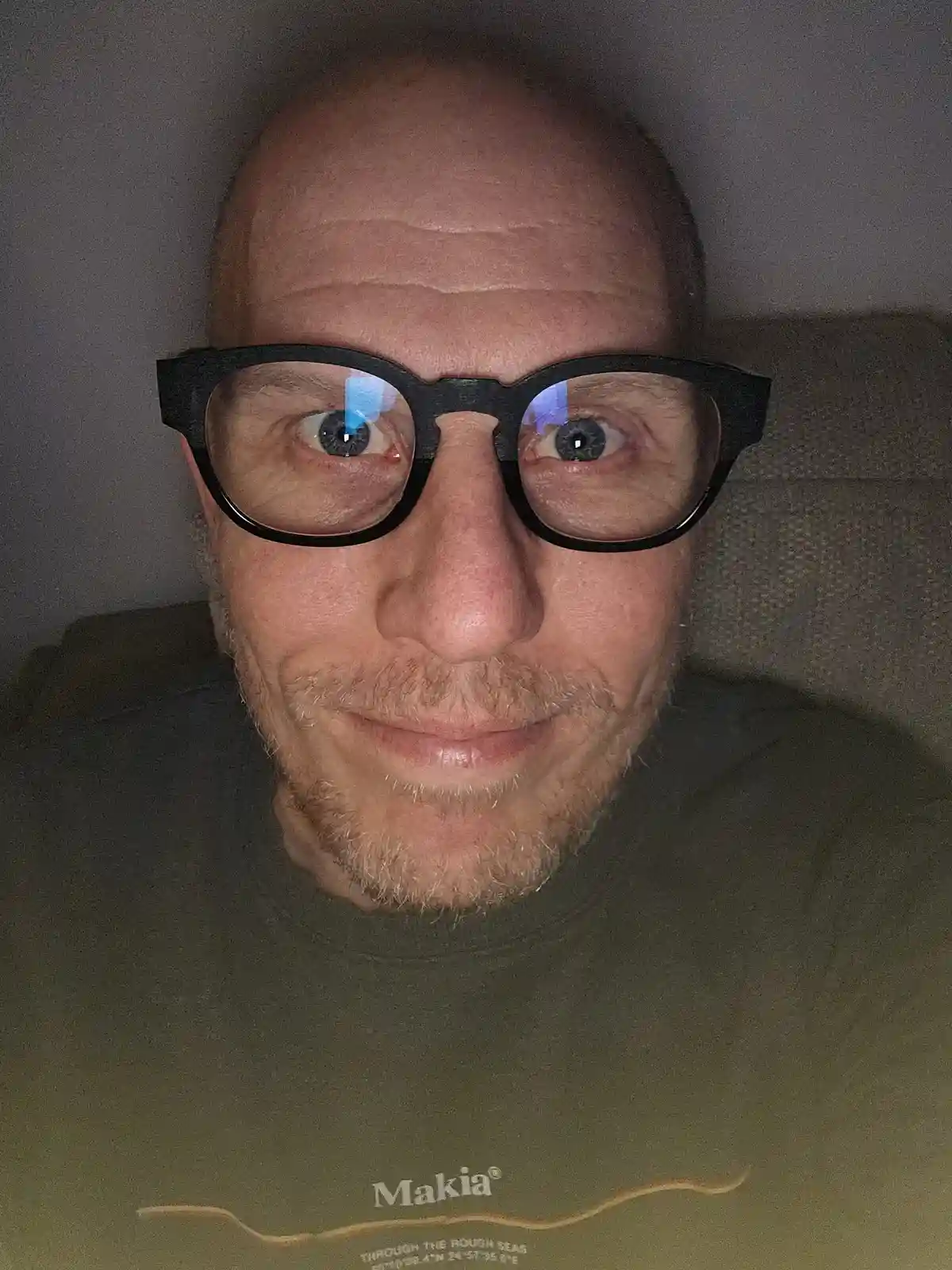 A close-up selfie of a bald man with a light beard wearing black-rimmed glasses, looking directly at the camera with wide…