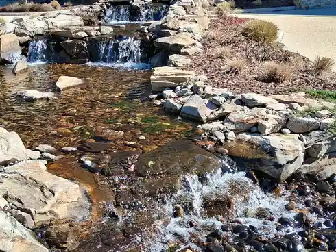 a serene outdoor scene featuring a man-made waterfall cascading over layered rocks into a shallow, clear stream. The water…