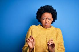 A woman with dark curly hair and a yellow sweater stands against a solid blue background, her face scrunched in an…