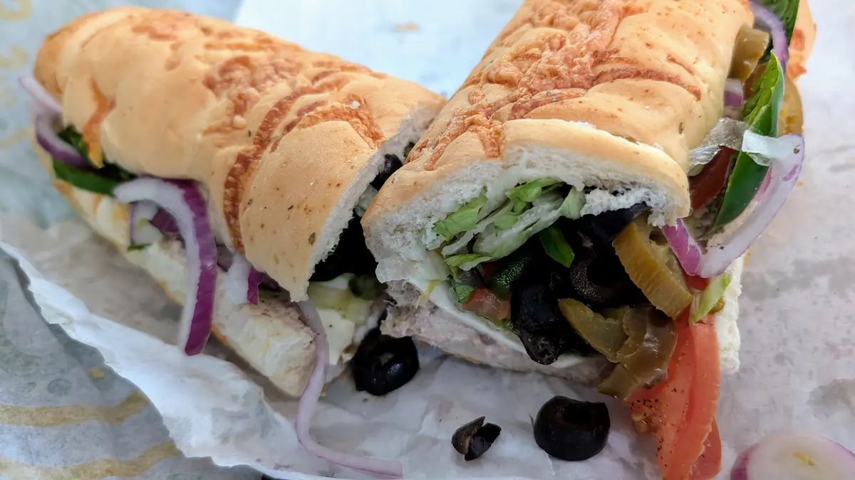 A close-up photo shows a freshly made sandwich cut in half, revealing layers of ingredients including lettuce, tomato, red…