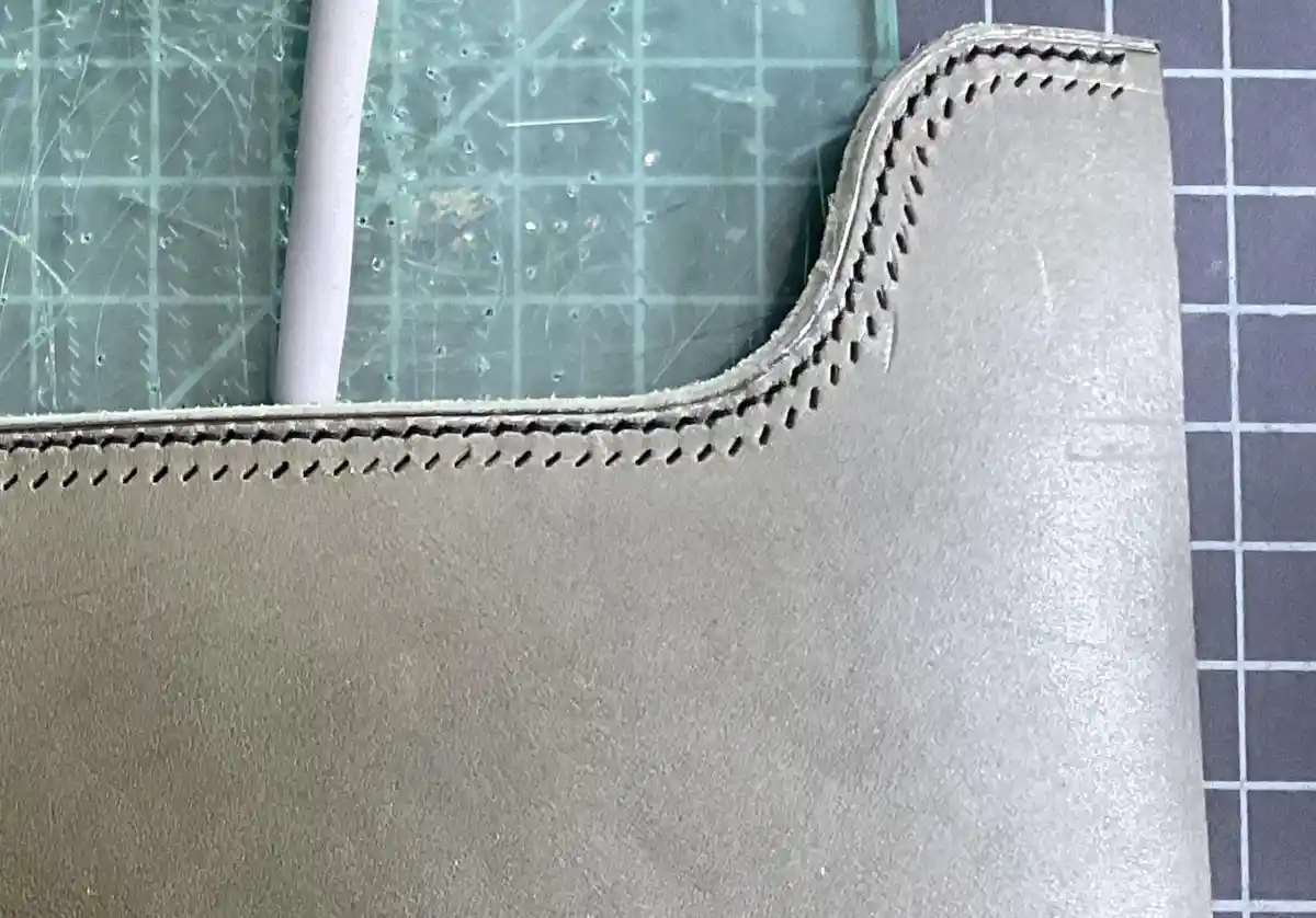 A close-up photo shows a curved edge of light gray leather with neat, evenly spaced stitching along its perimeter, resting…