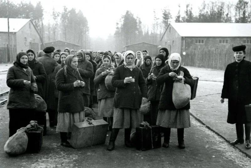 A black-and-white historical photograph shows a group of women in winter coats and headscarves standing in a line outdoors…