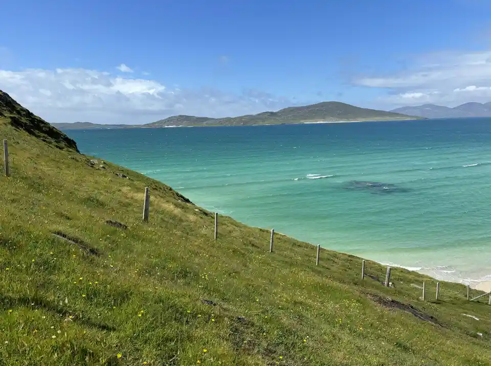 A scenic photo of a grassy coastal hillside overlooking turquoise ocean water with distant islands under a partly cloudy…