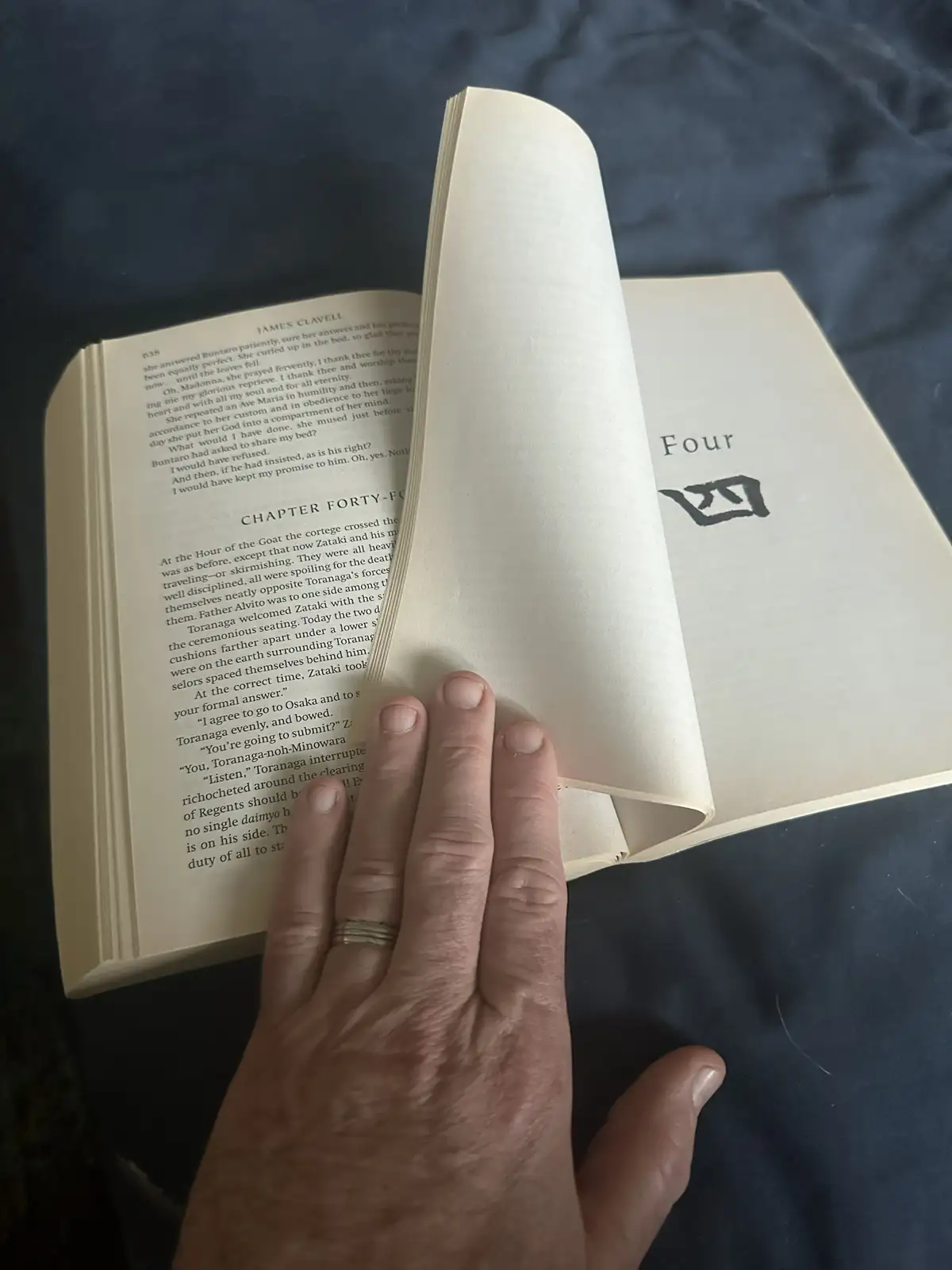 A hand rests on an open book, showing a page with text from James Clavell's novel. The right page displays the chapter…
