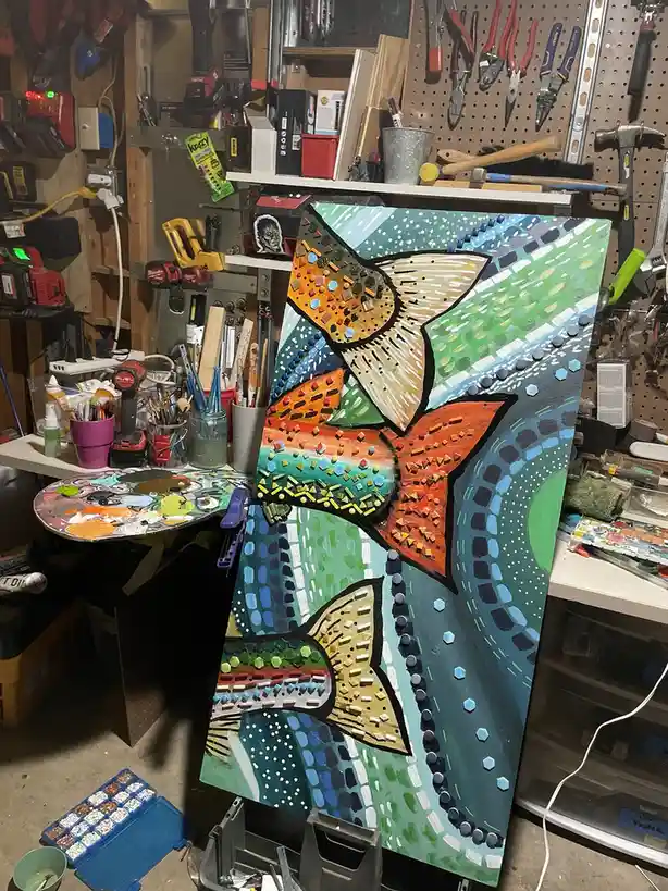 A colorful, textured mosaic-style painting of three fish is propped up in a cluttered artist's workshop filled with tools…