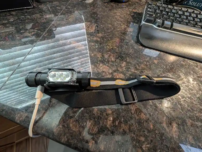 A black headlamp with a visible charging cable plugged into it lies on a dark granite desk next to a keyboard. The headlamp…