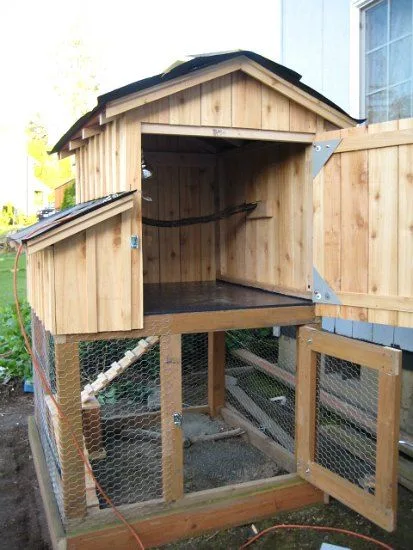 A wooden chicken coop with a slanted roof and wire mesh enclosure is shown outdoors, its main door and side gate open. The…