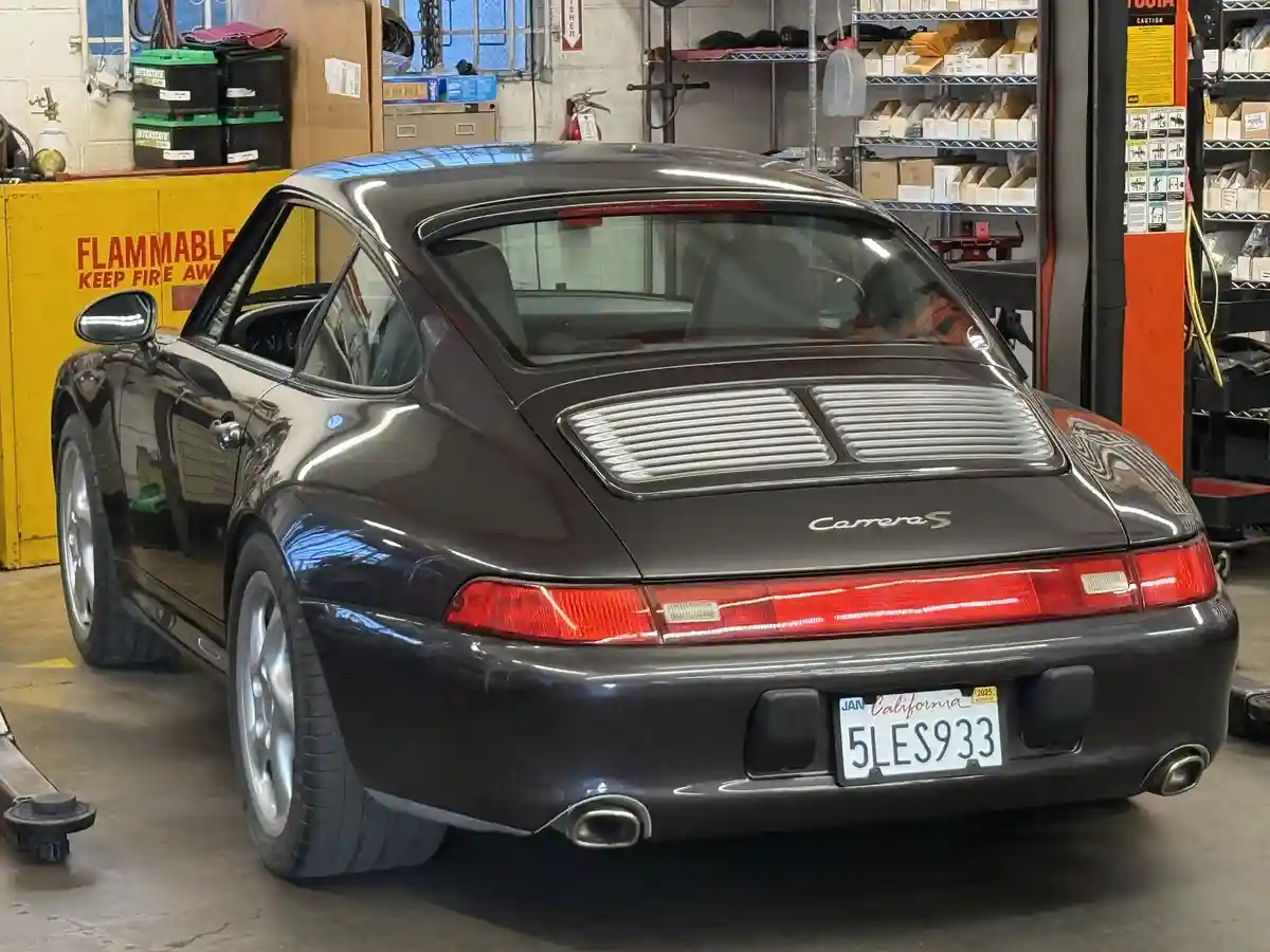 A dark gray Porsche 911 Carrera S is parked in a garage with its rear facing the camera, showing the California license…