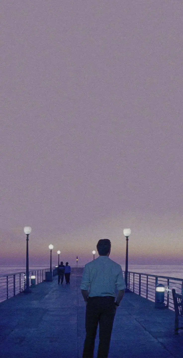 A man stands alone on a dimly lit pier at dusk, looking out over the water, with two other people walking away in the…