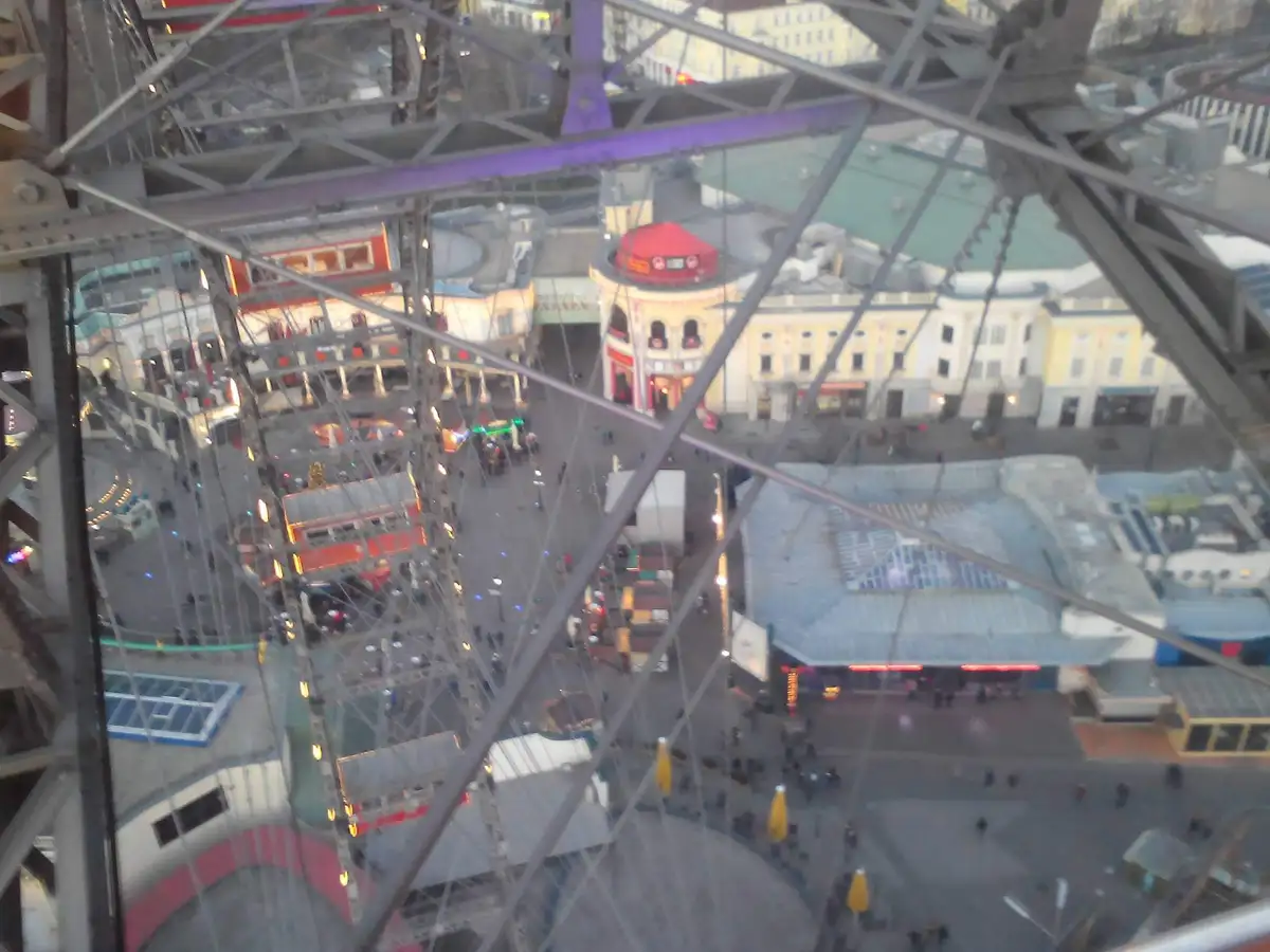 A blurry photo taken from inside a Ferris wheel cabin, looking down through the metal support structure at a crowded…