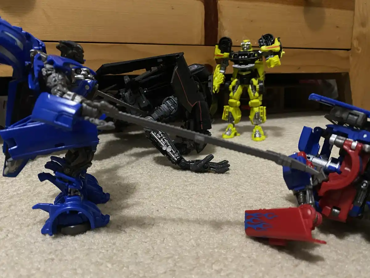 A blue Transformer toy holds a long sword, seemingly attacking a red and blue Transformer toy lying on its back on a beige…