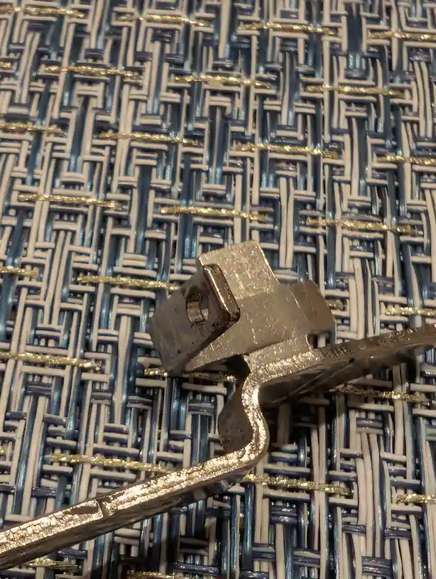 A close-up photo shows a bent, broken metal object, possibly a clip or latch, resting on a woven blue and white textured…