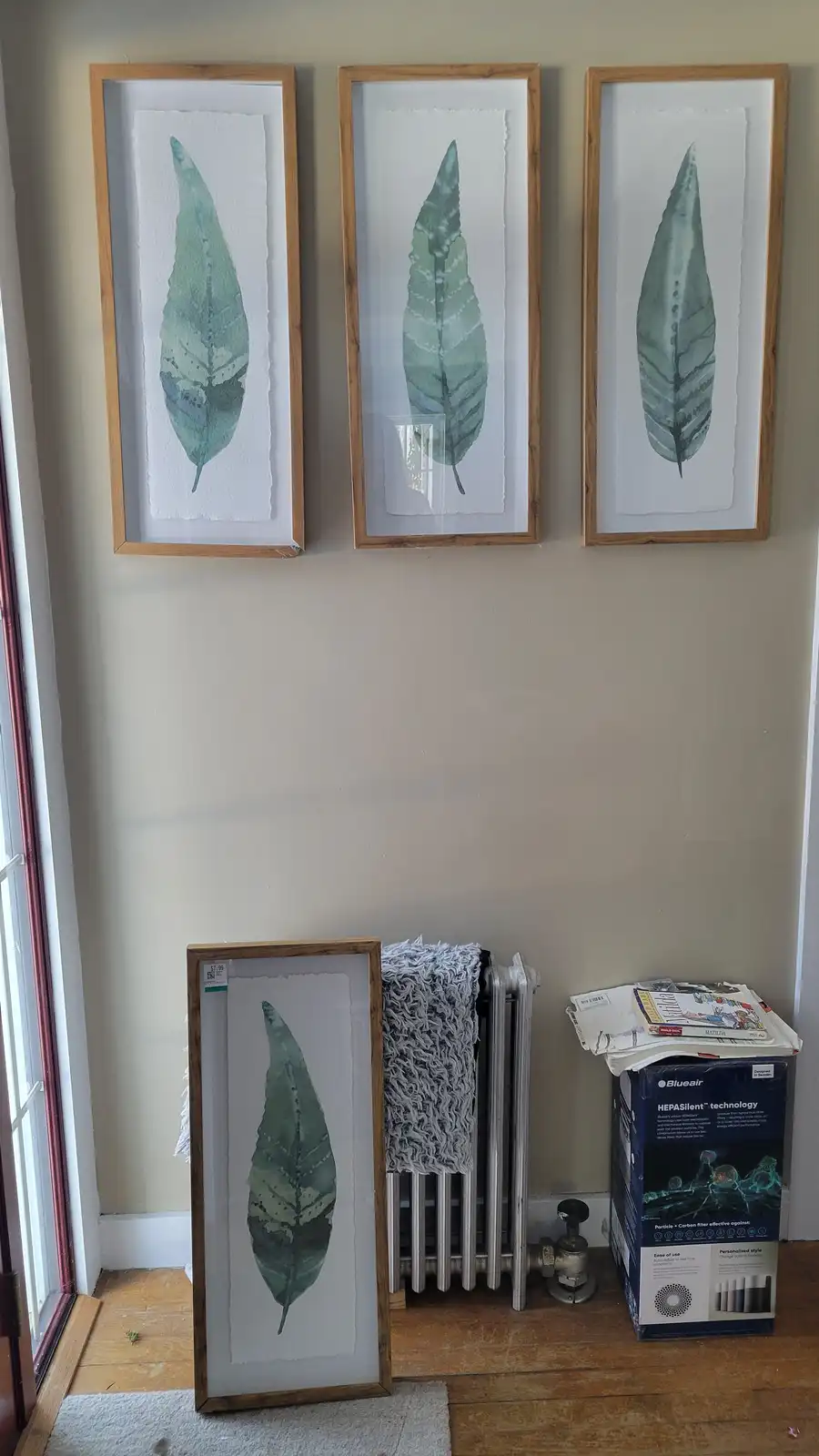Three framed leaf prints hang on a beige wall, while a fourth identical framed leaf print stands upright on the floor next…