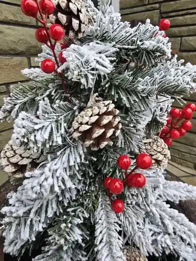 A close-up of a festive, artificial Christmas tree with green branches dusted in white faux snow, adorned with shiny red…