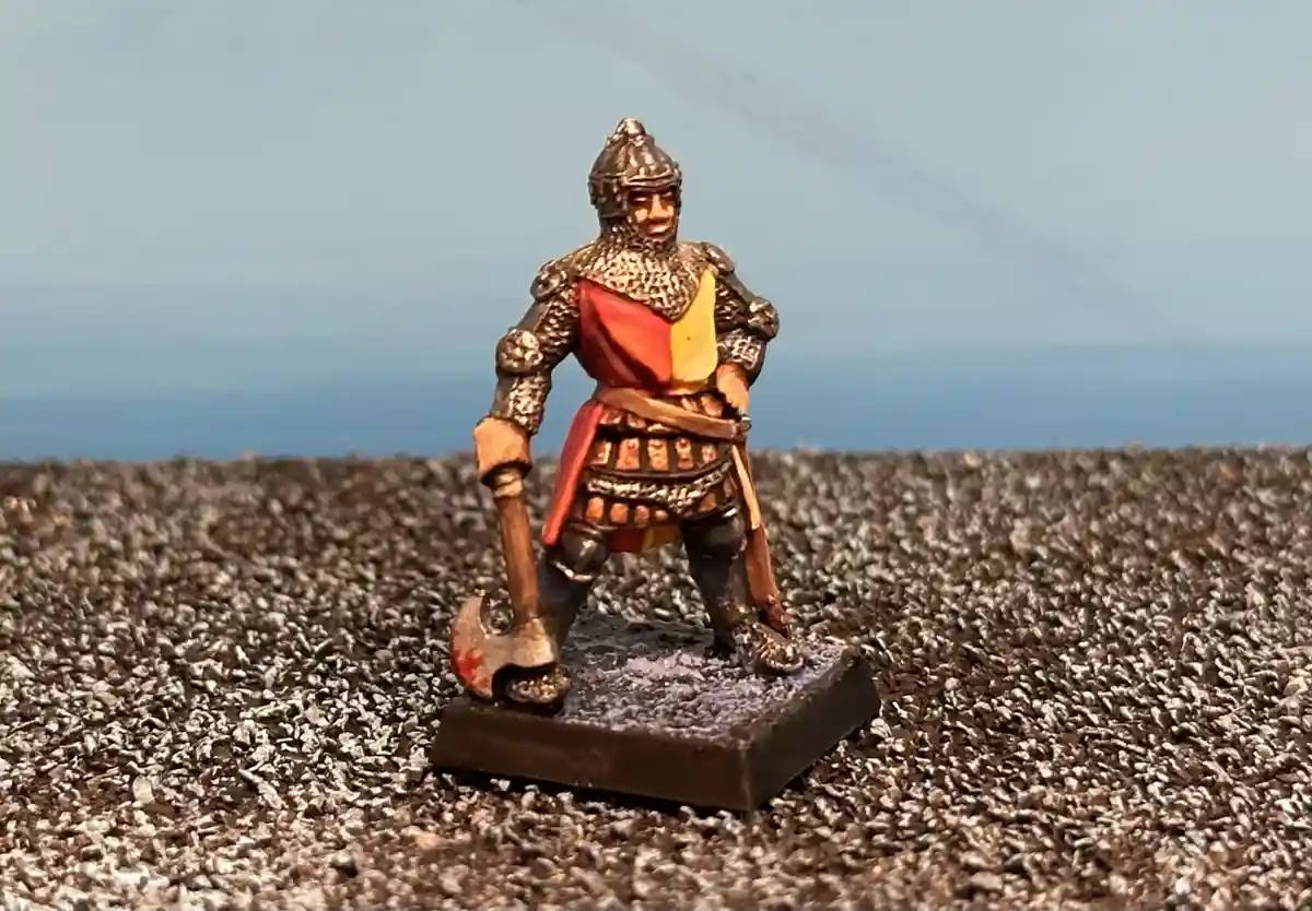 A painted miniature figurine of a medieval knight in full armor, holding an axe, stands on a textured base against a blurred…