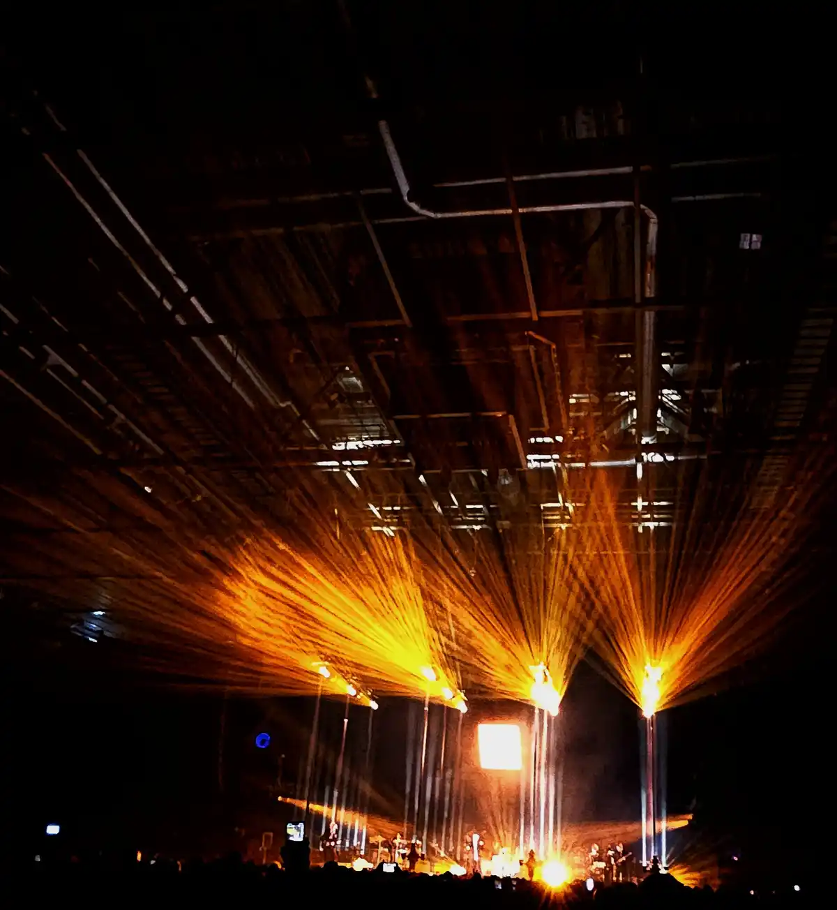 A dark concert venue with bright orange spotlights radiating from the stage upward toward the ceiling, illuminating dust…