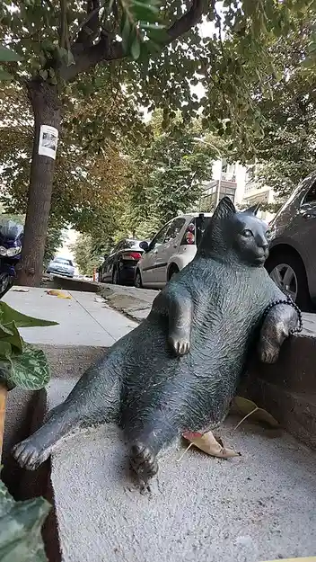 A bronze statue of an overweight cat lounging lazily on a concrete ledge outdoors, surrounded by trees and parked cars. The…