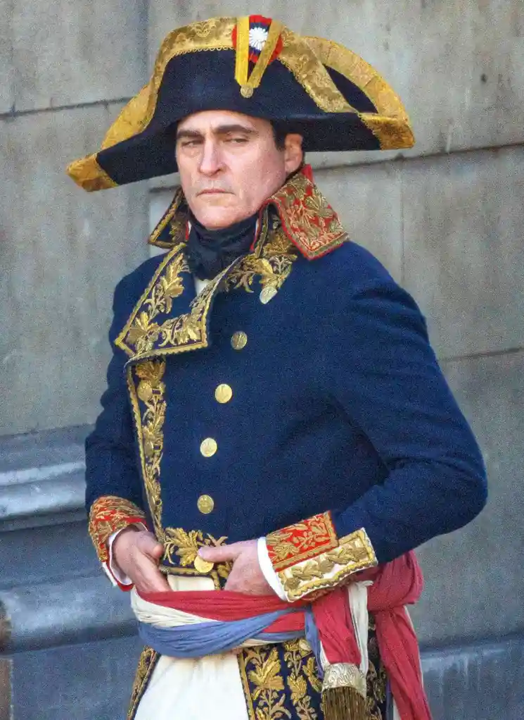A man dressed in an ornate, historical French military uniform with gold trim, a bicorne hat, and a stern expression, stands…