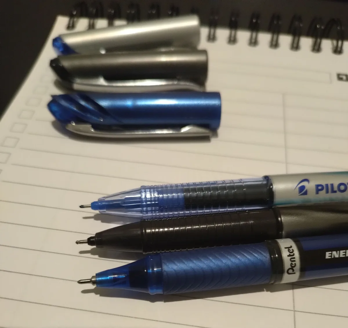 Several ballpoint pens, some with caps on and some uncapped, are arranged on a lined spiral notebook. The pens vary in color…