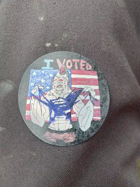 A hand-drawn, circular 'I VOTED' sticker is pinned to a dark gray fabric surface. The sticker features a cartoonish…