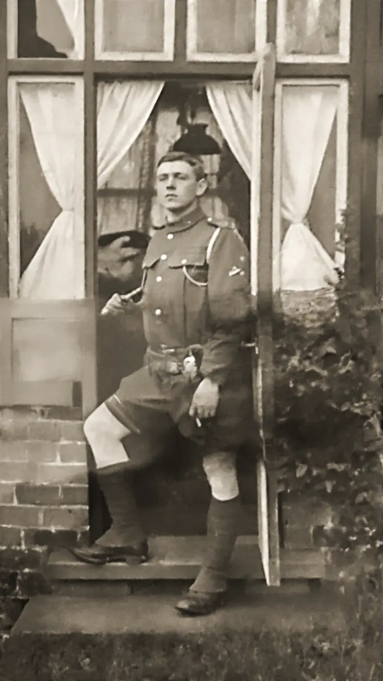 A sepia-toned historical photograph shows a young man in early 20th-century military uniform standing on a doorstep, leaning…