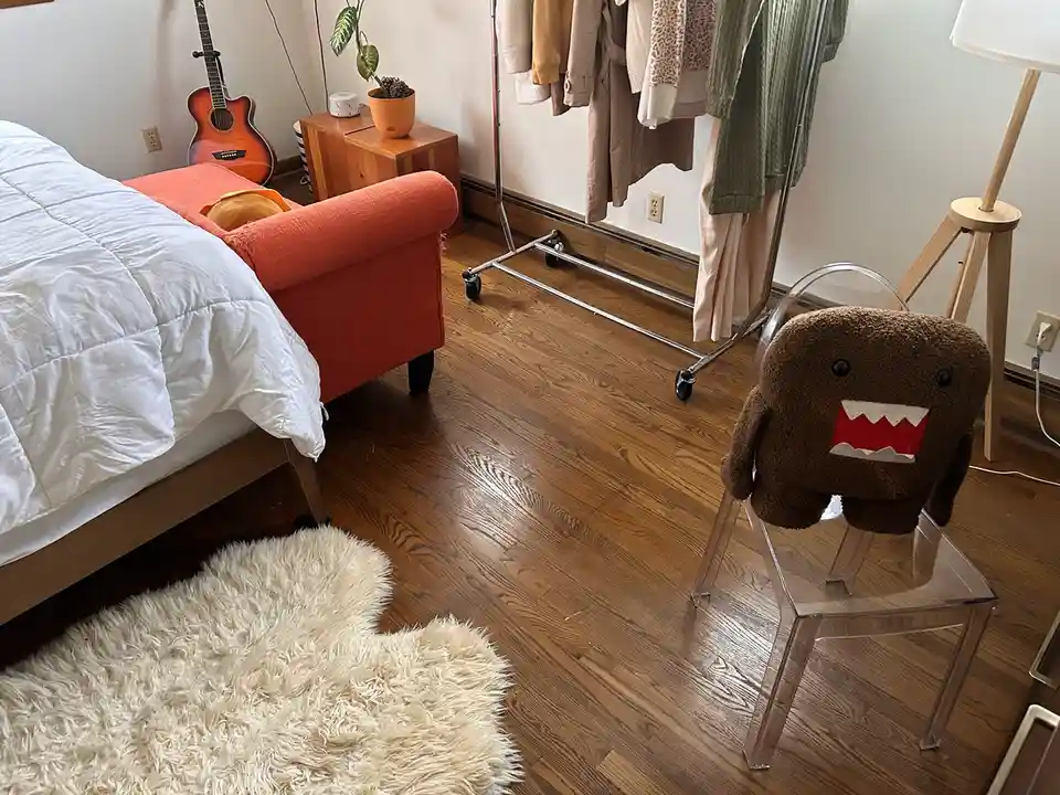 A brown, fuzzy Domo plush toy with a wide red mouth sits on a clear acrylic chair in a bedroom. The room contains a bed with…