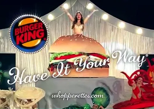 A woman stands atop a giant Burger King Whopper burger on a stage with white curtains and spotlights. The Burger King logo…