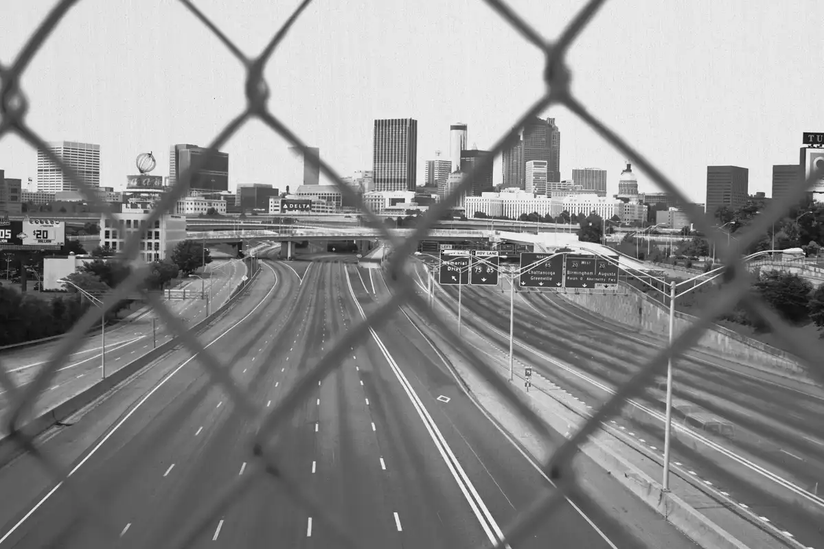 A black-and-white photo shows an empty highway with a city skyline in the background, viewed through a chain-link fence…