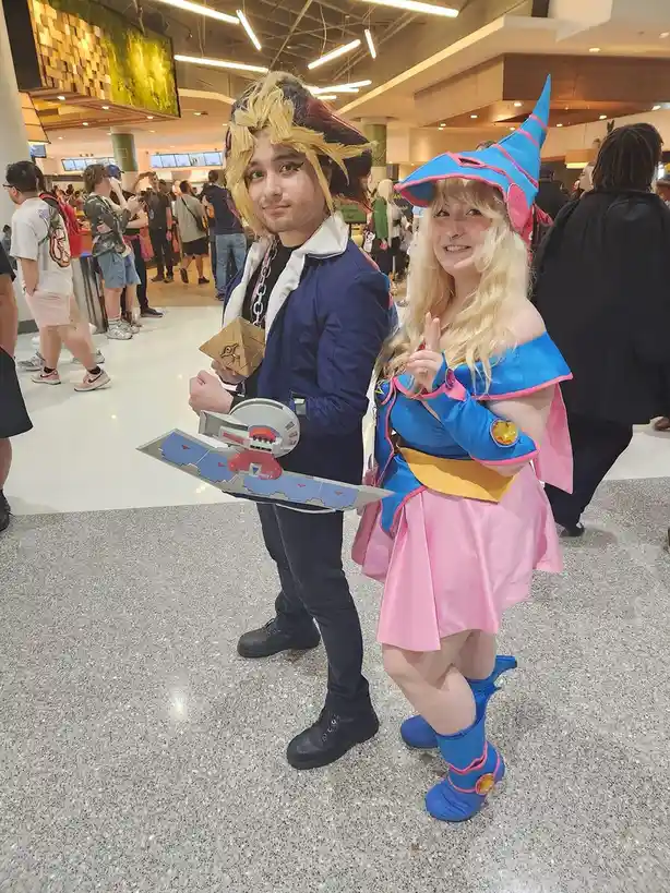 Two cosplayers dressed as Yugi Muto and Yami Yugi from Yu-Gi-Oh! pose together at a convention. The male cosplayer wears a…