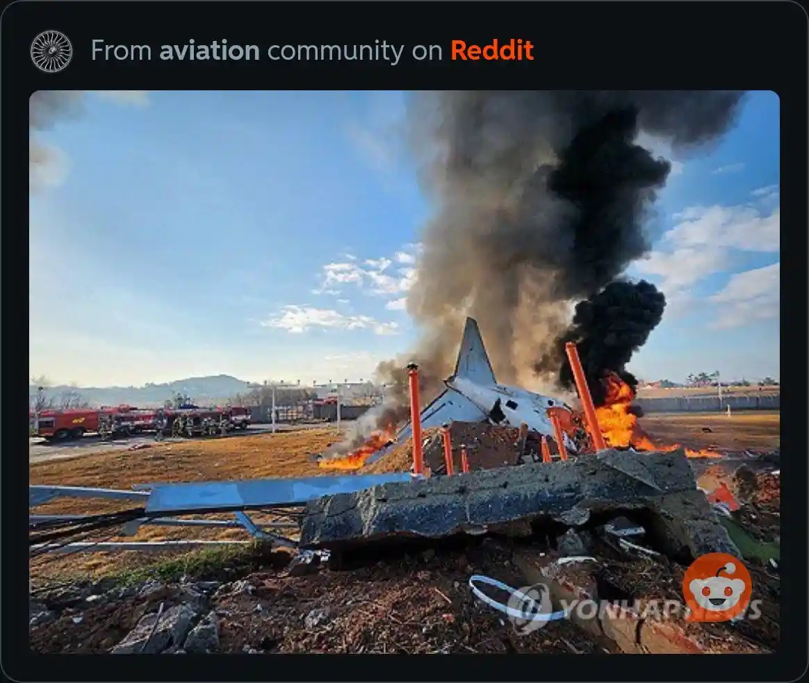 A crashed airplane is on fire with thick black smoke rising against a blue sky, surrounded by emergency vehicles and debris…