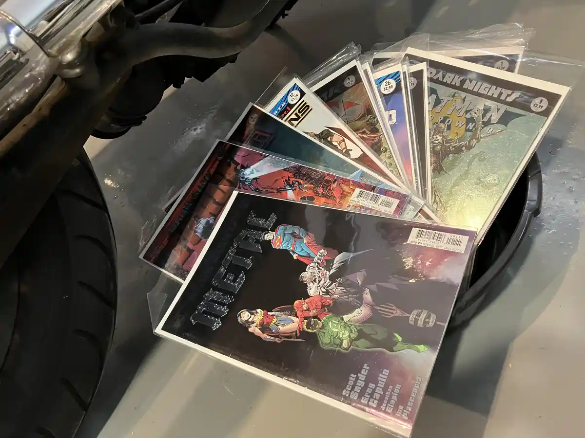 A fanned-out pile of DC Comics, including 'Dark Nights: Batman' and 'Metal', rests on a car's tire and wheel well. The…