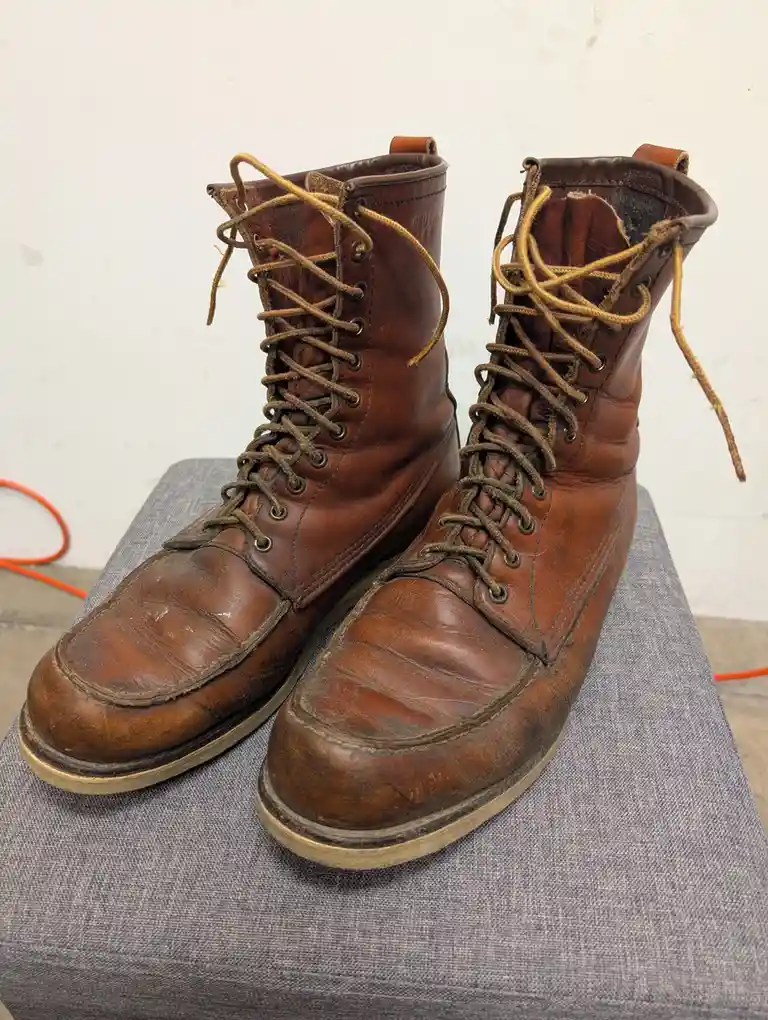 A pair of well-worn, brown leather work boots with frayed laces sit on a gray fabric surface. The boots show significant…