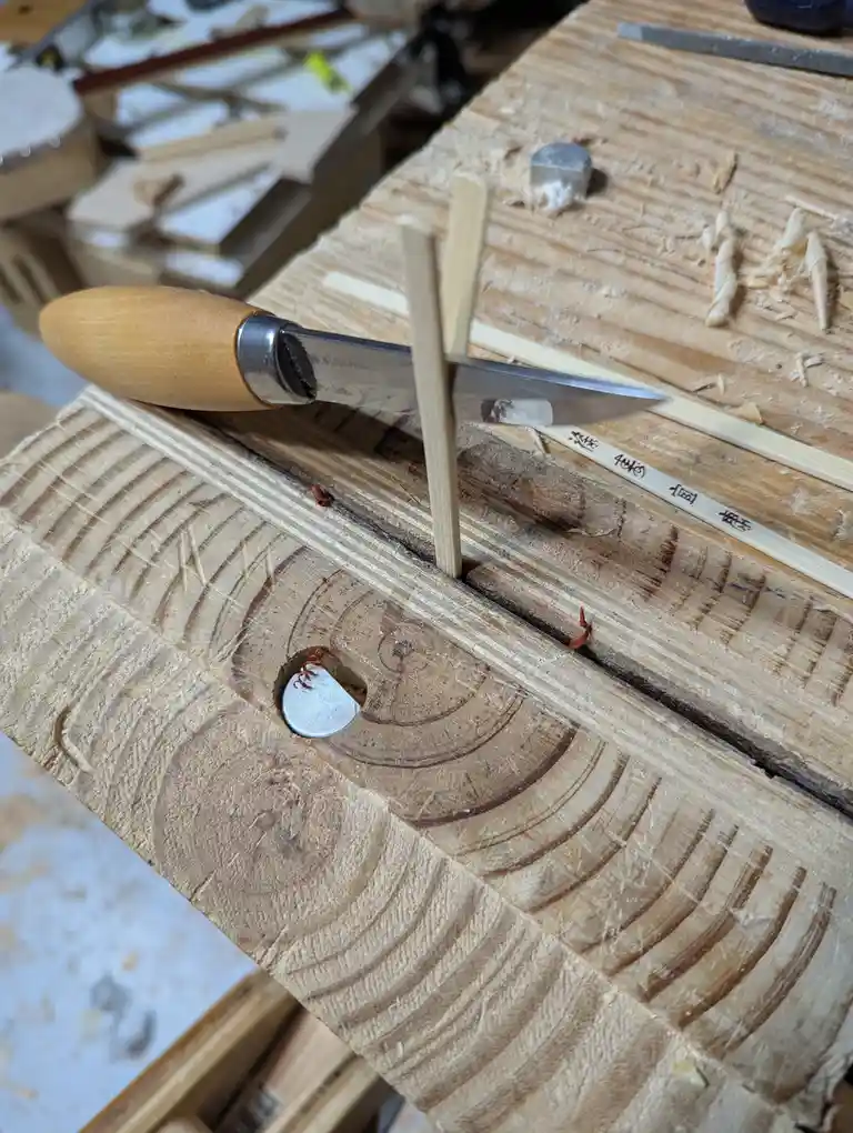 A close-up photo of a woodworking bench with a carving knife, wooden sticks, and wood shavings. A small, round metal disc…