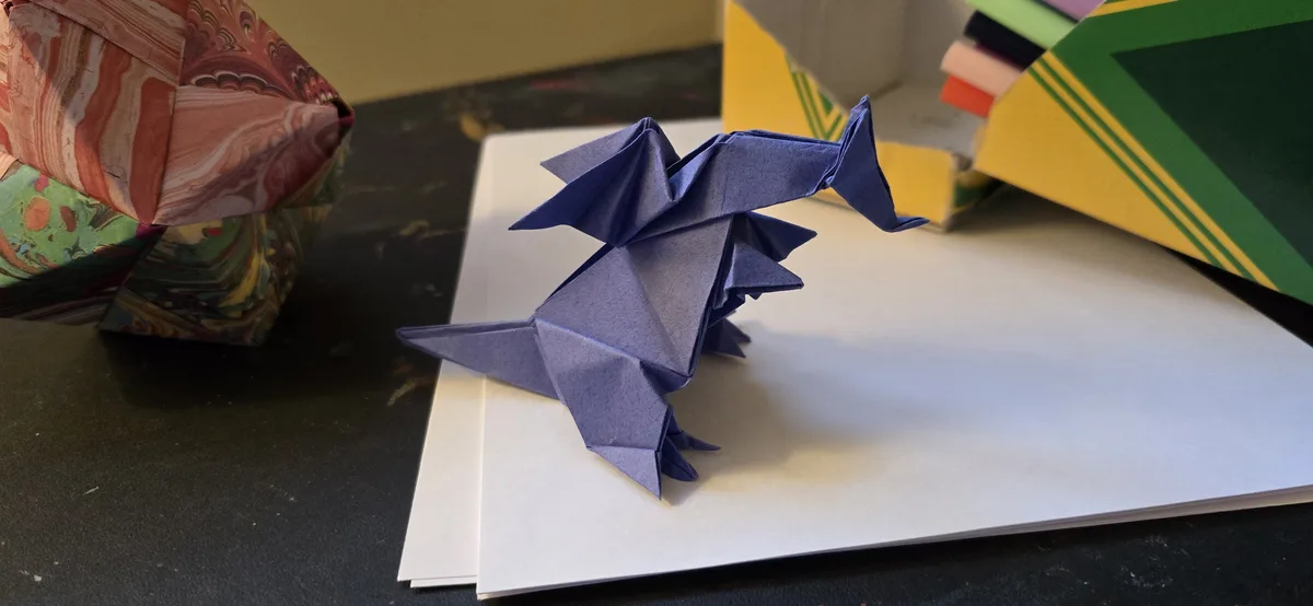 A blue origami dragon sits on a stack of white paper, surrounded by colorful folded paper stacks. The overlaid text reads 'i…