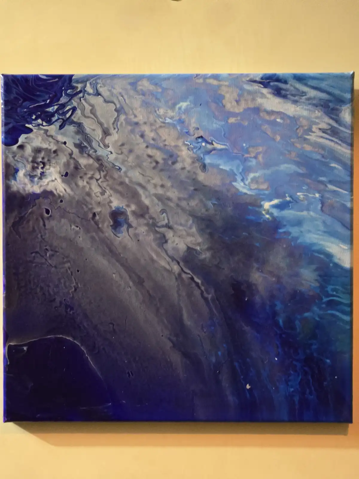 A square canvas painting with swirling blue, purple, and gray abstract patterns resembling a galaxy or nebula, hanging on a…