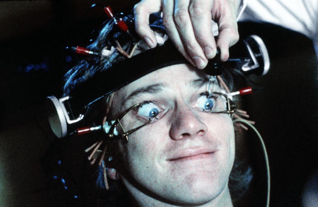 A close-up of a young man with wide, startled eyes, wearing a complex head-mounted apparatus with wires and sensors, while a…