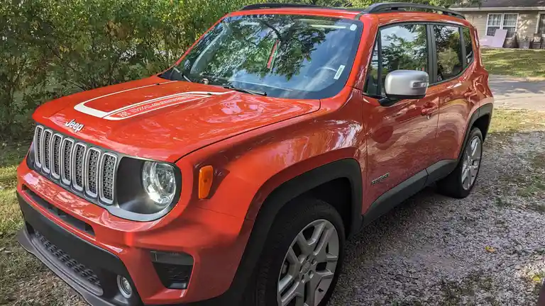 A red Jeep Renegade SUV is parked on a gravel driveway with green bushes and a house in the background. The car's hood has a…