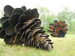 Two large, dark, metallic pinecone sculptures sit on grass, one large in the foreground and one smaller in the background…