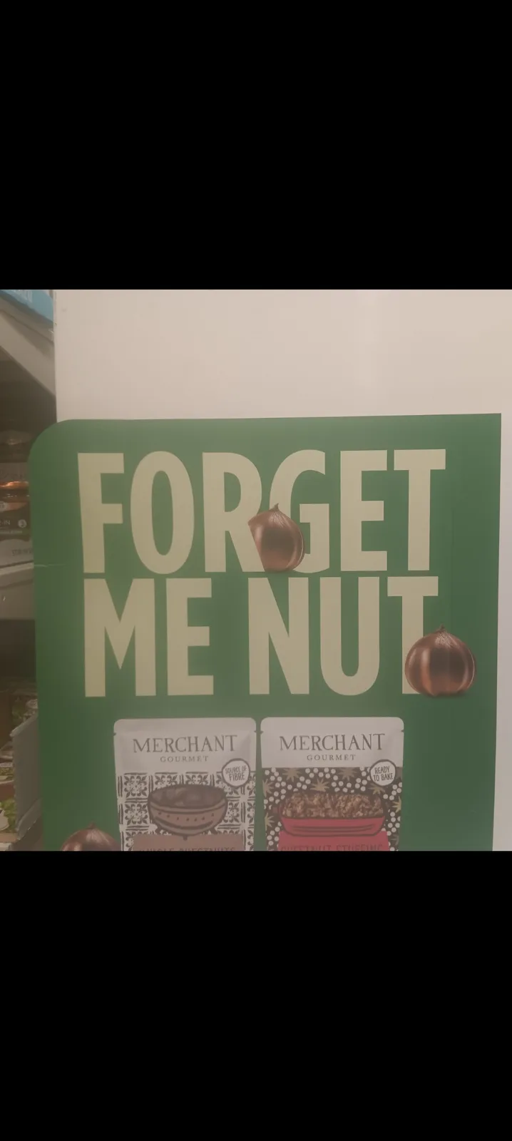 A green store advertisement sign for 'Merchant Gourmet' chestnut products features the text 'FORGET ME NUT' with chestnuts…
