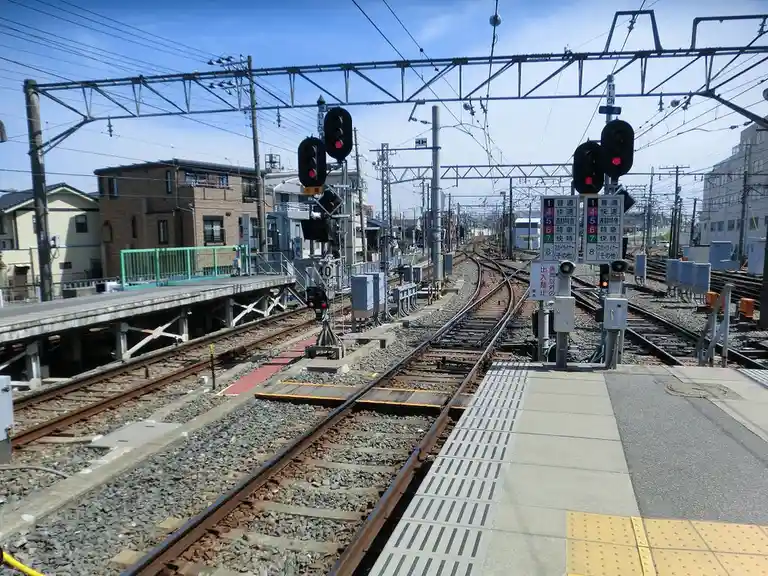 A photo of a Japanese railway station platform with multiple tracks, overhead wires, and signal lights showing red. A sign…