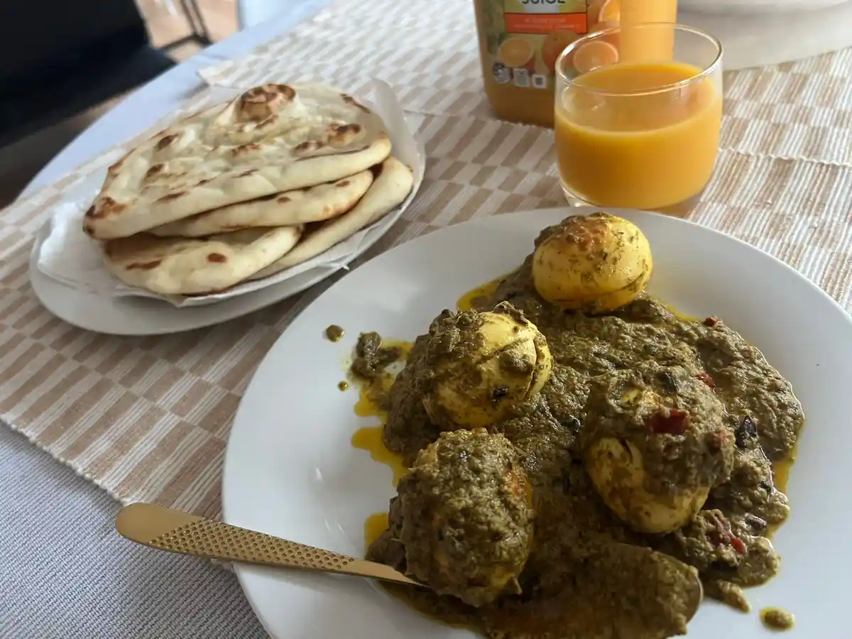 A plate of Indian-style curry with boiled eggs and a side of naan bread is served alongside a glass of orange juice on a…