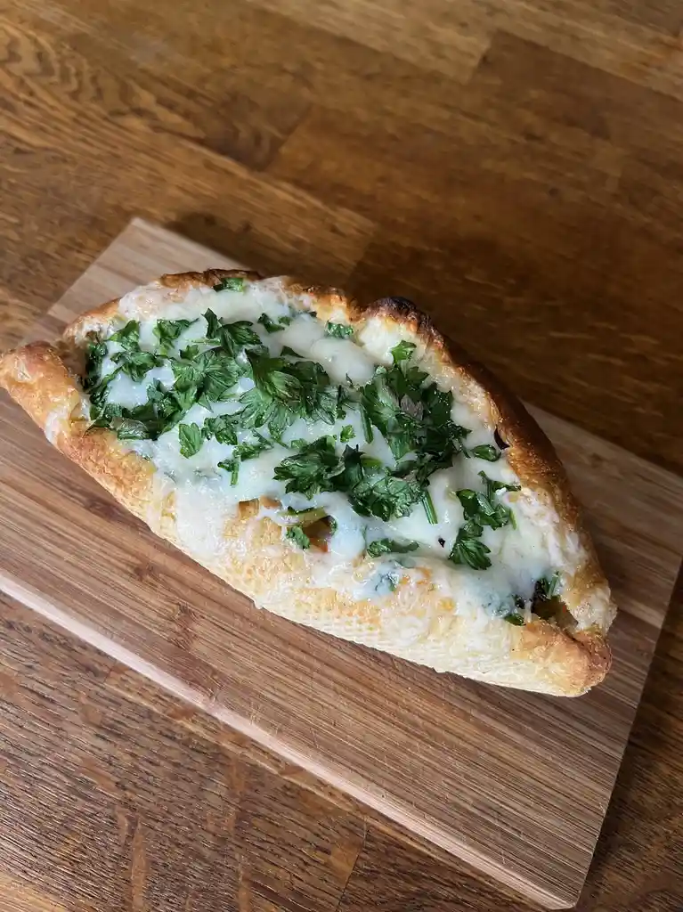 A close-up photo shows a single slice of toasted bread, likely a baguette, filled with melted white cheese and topped with…