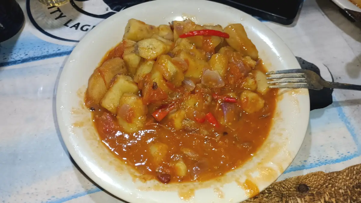 A white plate holds a dish of stewed chunks of food in a thick orange-red sauce, garnished with red chili and onions, with a…