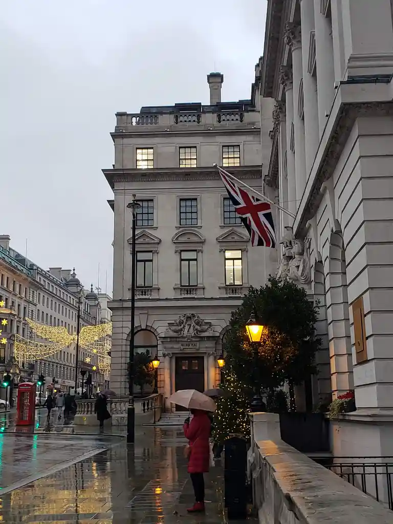 A rainy street scene in London features a person in a red coat holding an umbrella, standing near a grand building with a…