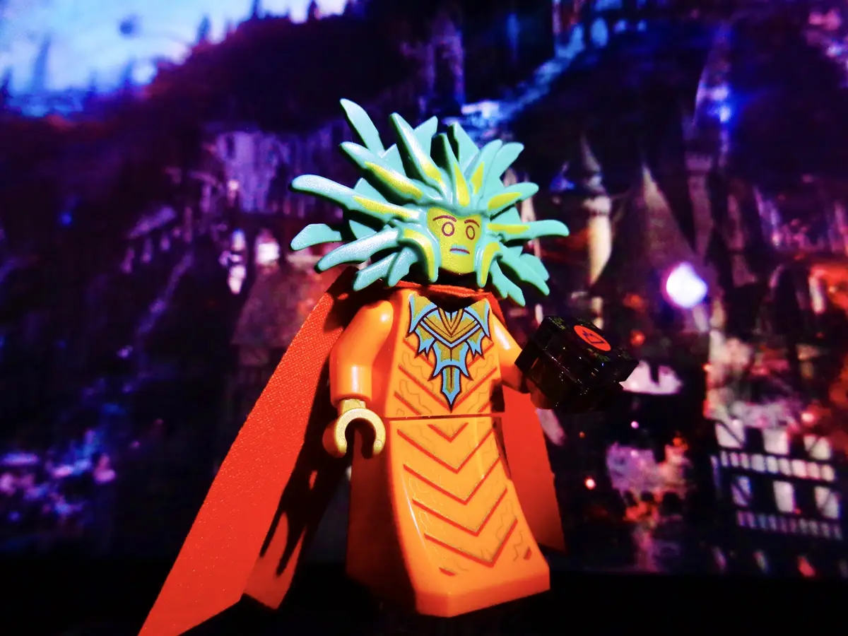 A LEGO minifigure with a spiky green and yellow headpiece, orange torso with blue and gold patterns, and a red cape stands…