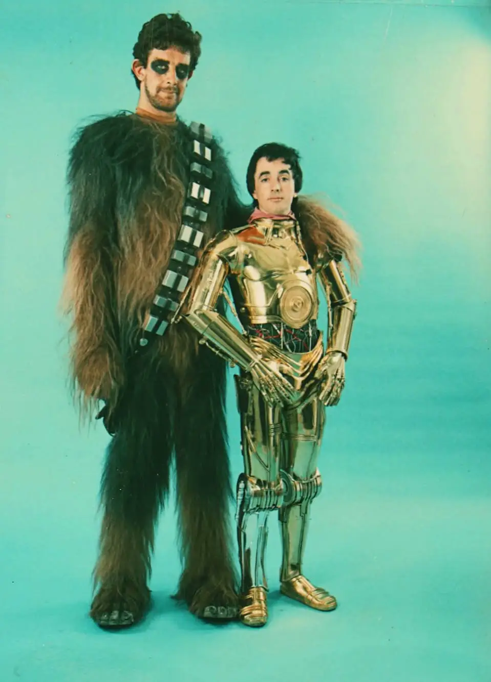Two men pose against a teal backdrop; one dressed as Chewbacca with fur and black eye makeup, the other in a gold C-3PO…
