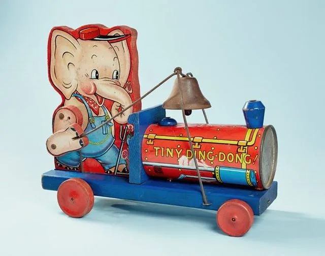 A vintage tin toy train with a cartoon elephant driver wearing overalls and a red hat, pulling a red and blue train car…