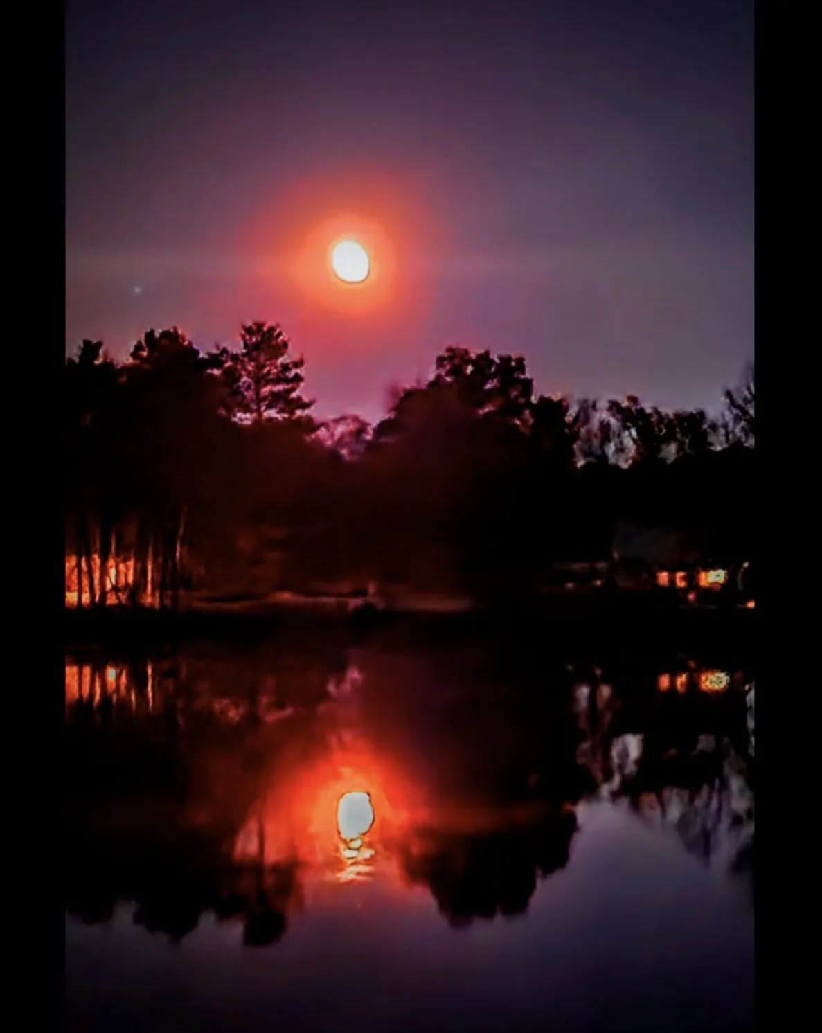 A serene night scene shows a bright, glowing moon above a dark treeline, reflected in a calm body of water below. The moon's…
